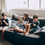 Three children on a gray sofa engrossed in digital devices, indoors.